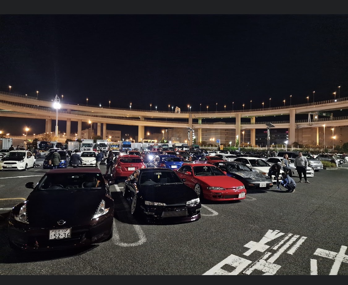 A nighttime car meet at Daikoku Parking Area, with rows of modified cars and highway overpasses lit up in the background